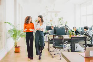 Two women walking side by side through an office, carrying lunch and t